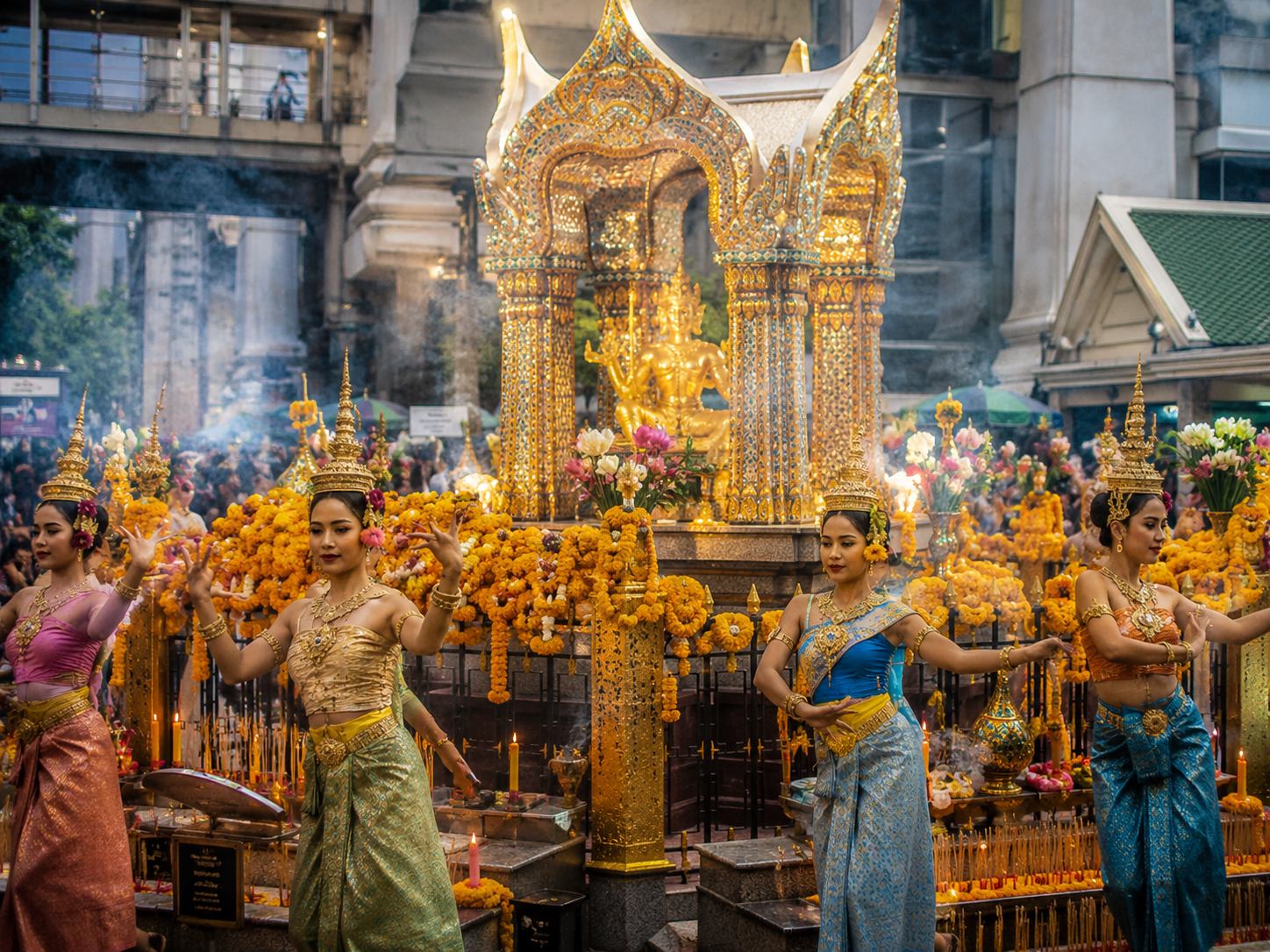 Devotees offering incense and flower garlands at the golden Erawan Shrine at Bangkok's Ratchaprasong intersection, with the Grand Hyatt Erawan hotel in the background